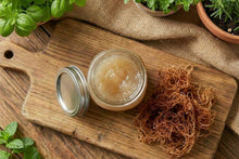 Jar of cream, seaweed, and herbs on a wooden board with plants in the background